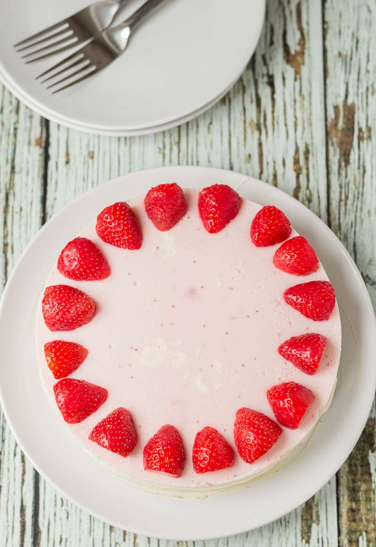 Birds eye view of a whole no bake strawberry mousse cheesecake decorated around the edge with halved strawberries. Serving plates and forks at the top.