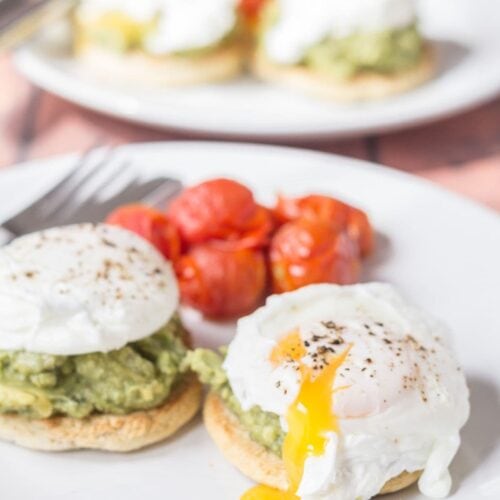 Two plates of smashed avocado and poached eggs on muffins, one in front of the other. Each plate served with cherry tomatoes and a knife and fork on.