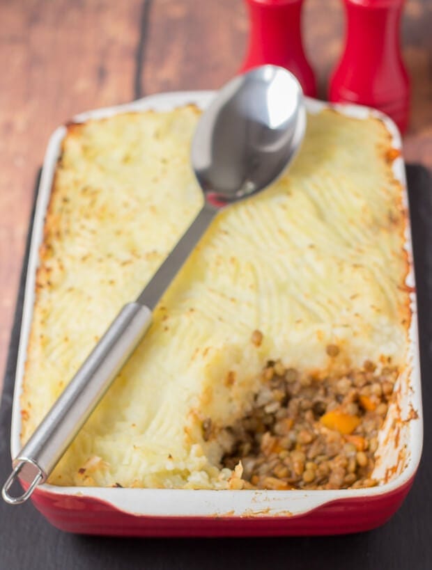 A casserole dish of cooked green lentil shepherd's pie with a portion removed. Serving spoon sitting over the top.