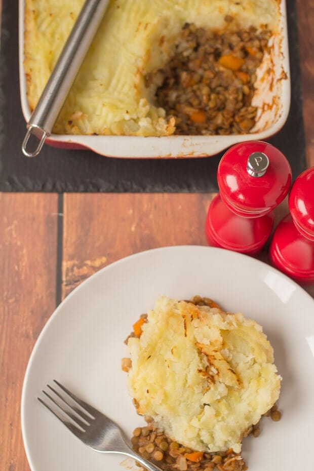 Birds eye view of a portion of green lentil shepherd's pie served on a plate at the bottom. Part of the cooked casserole dish at the top with salt and pepper cellars in between. 