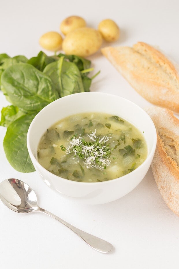 A bowl of potato and spinach soup with a soup spoon, spinach leaves and potatoes arranged around and two halves of a baguette.