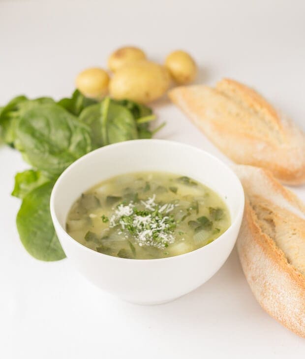A bowl of potato and spinach soup with spinach leaves and potatoes arranged around and two halves of a baguette.