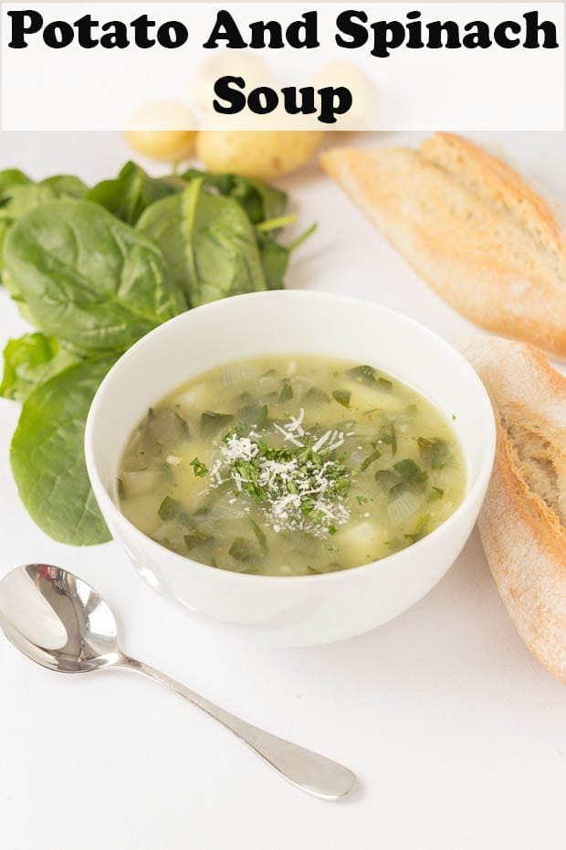 A bowl of potato and spinach soup with a spoon beside the bowl and bread and spinach surrounding it.