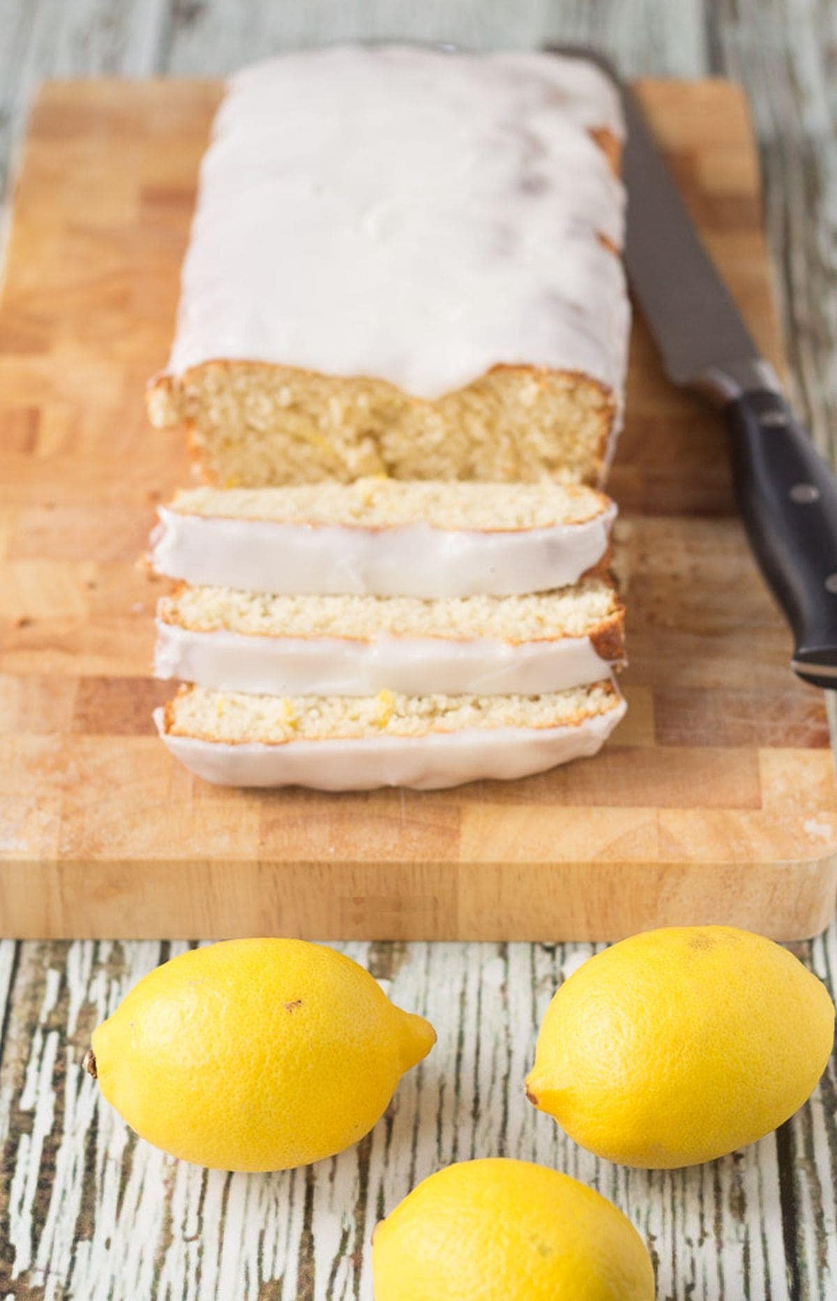 A lighter lemon loaf cake on a chopping board facing forward with three slices cut off. Three lemons in front as decoaration and a carving knife to the left hand side.