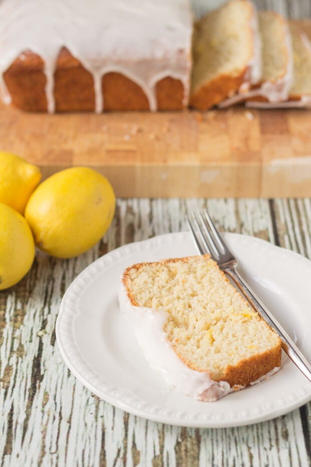 A slice of lighter lemon loaf cake on a plate with a fork. The rest of the loaf cake on a chopping board in the background with three lemons in between.
