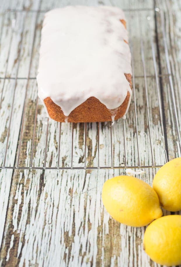 An iced lighter lemon loaf cake on a wire baking rack. Three whole lemons in the foreground.