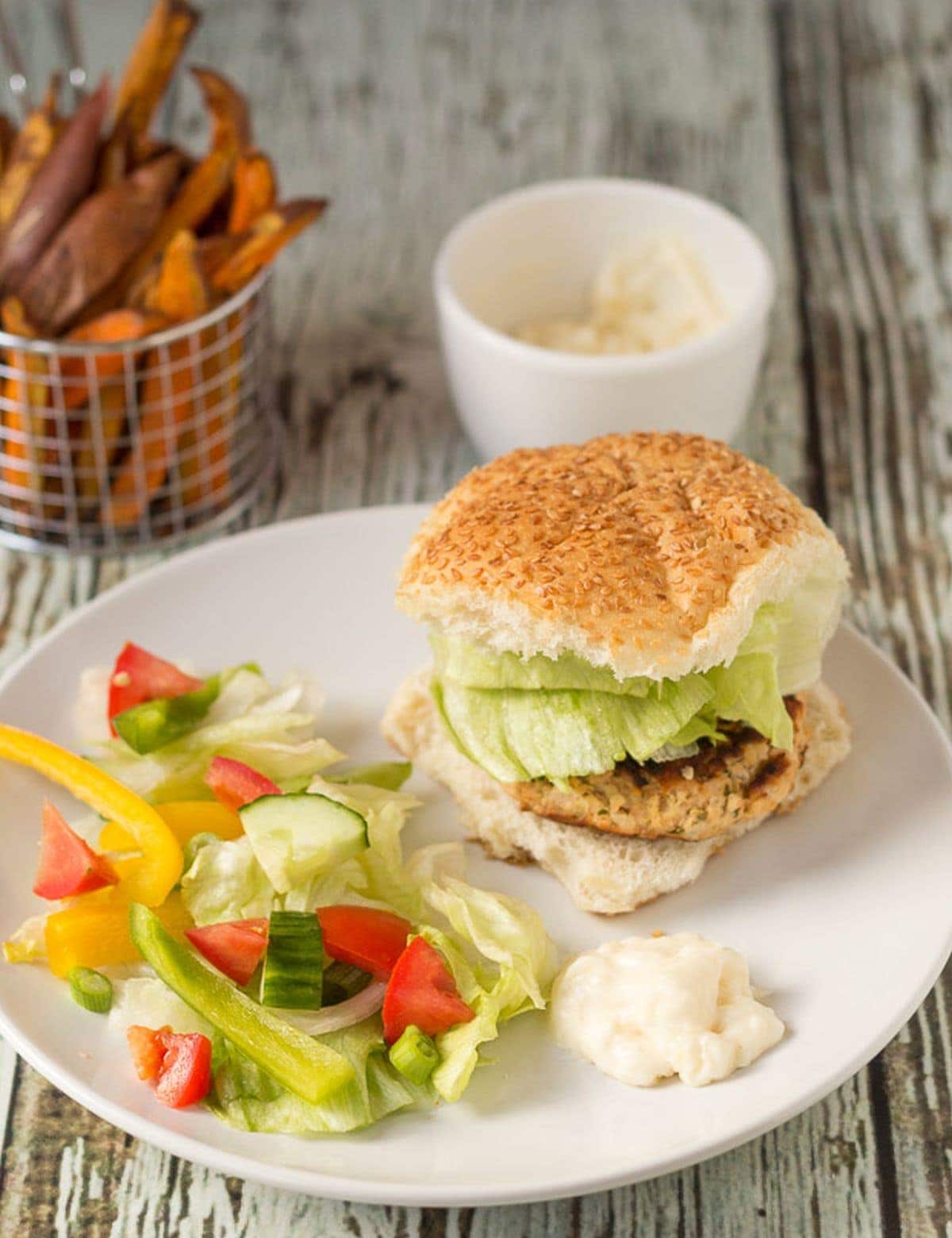A quick healthy salmon burger served in a bun on a plate with salad. Pot of mayonnaise and basket of sweet potato fries in the background.