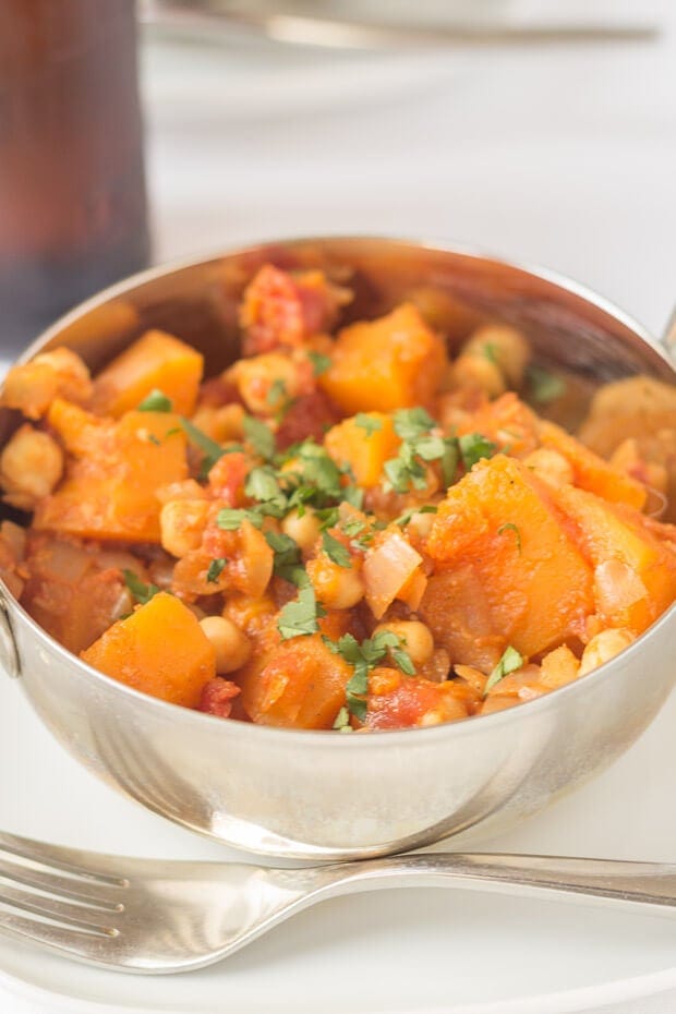 Close up of a balti dish with chickpea and butternut squash curry in, sitting on a plate with a fork to the side. A bottle of lager in the background.