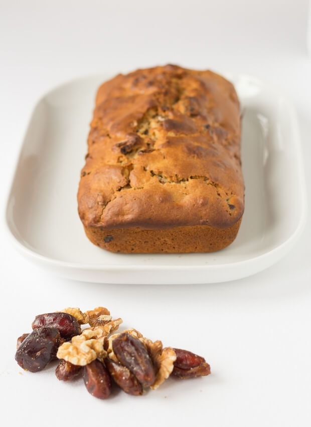 Date and walnut loaf on a white serving platter. A pile of date and walnuts in front.