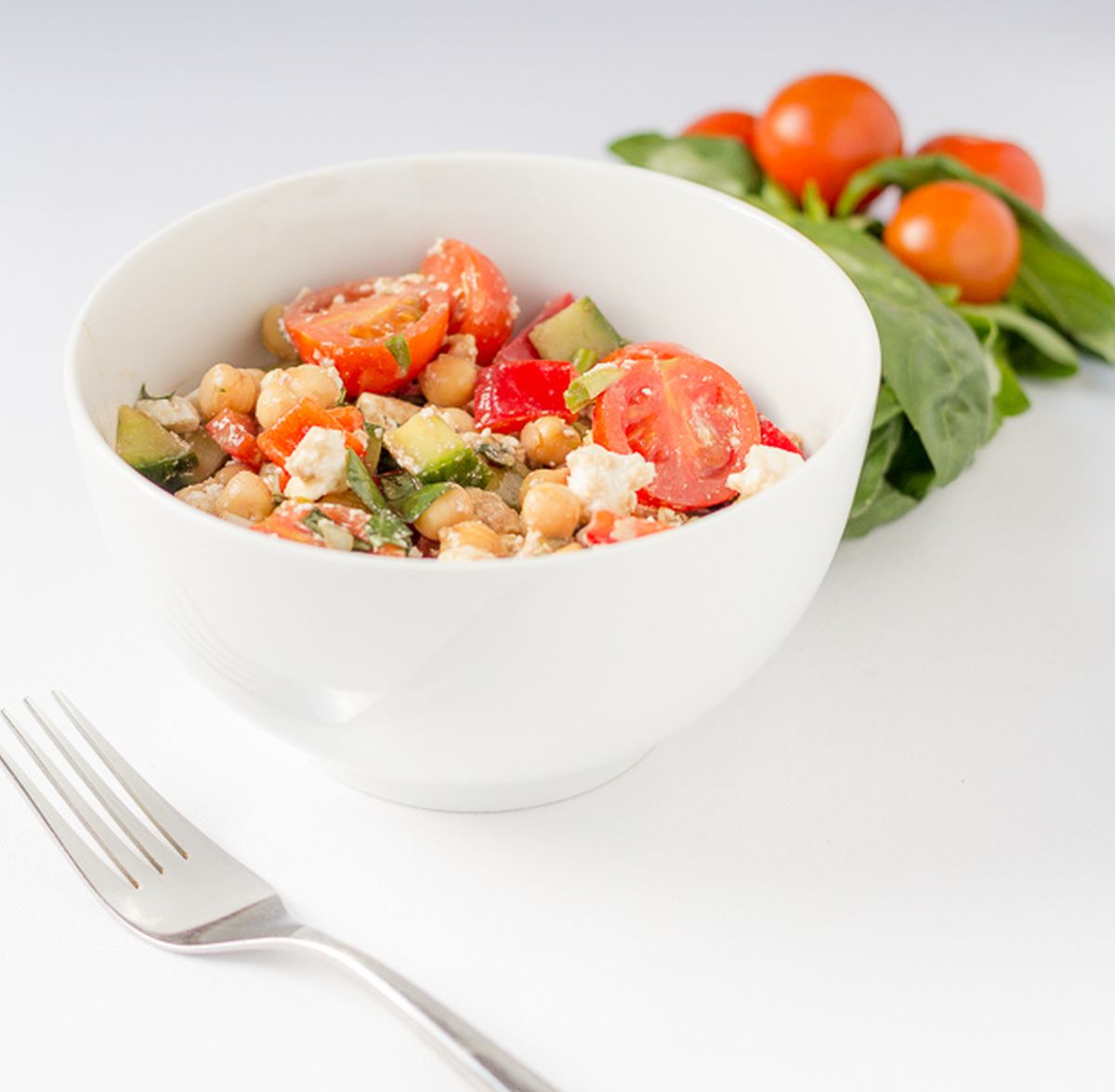 A bowl of Mediterranean chickpea salad with a fork beside. Basil leaves and cherry tomatoes in the background.