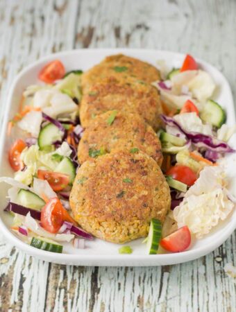 Four quick healthy chickpea cakes arranged on a platter dish surrounded by a salad of lettuce, cucumber and sliced tomatoes.