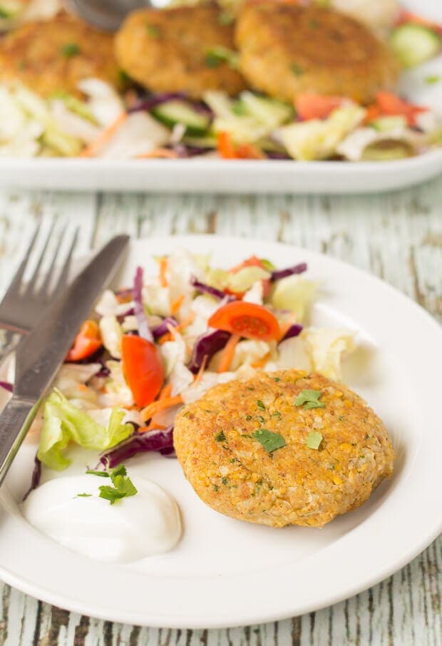 A quick healthy chickpea cake served on a plate with some salad and a knife and fork to the side. Rest of the three chickpea cakes on a platter dish in the background.