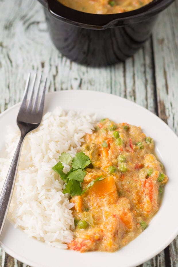 Close up view of a plate of slow cooker sweet potato and vegetable curry served with rice and a fork to the side. Rest of the recipe in slow cooker dish at the top.