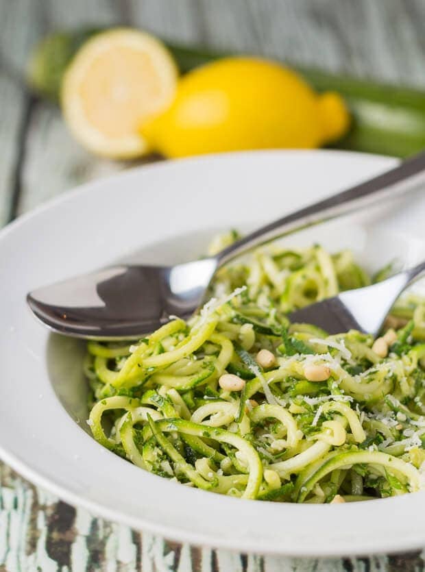 Close up view of a bowl of spiralized kale pesto pasta with a fork and spoon in. Two lemons and a courgette in the background.