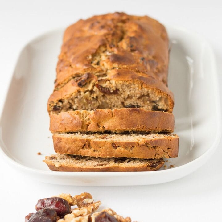 Date and walnut loaf on a white serving platter facing forward with three slices cut off. A pile of date and walnuts in front.