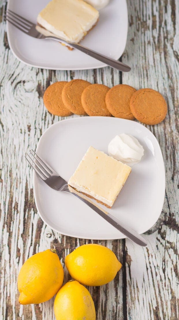 Two plates with a lemon cheesecake square served on a plate with a fork to the side. Biscuits and lemons around the plates as decoration.