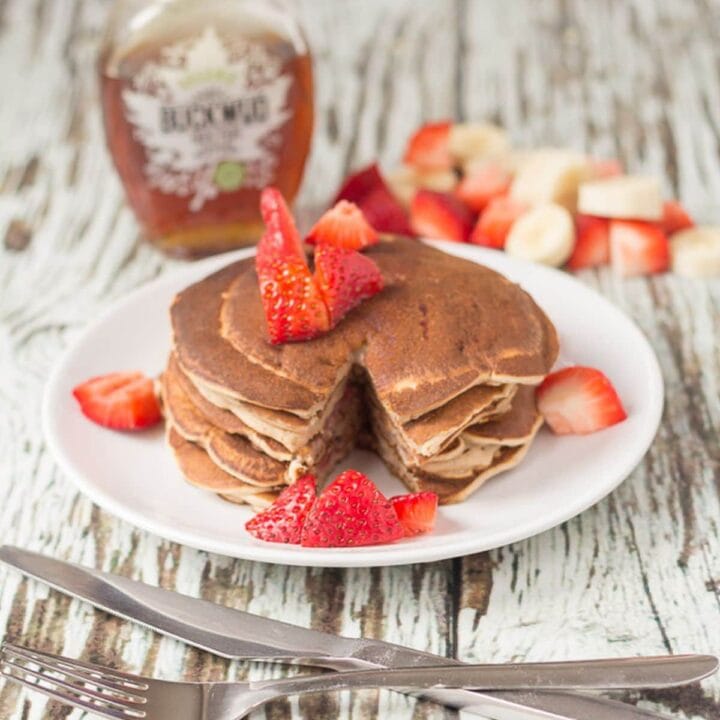 A stack of strawberry banana valentines pancakes on a plate topped with chopped strawberries. Knife and fork to the front. A bottle of maple syrup in the background.