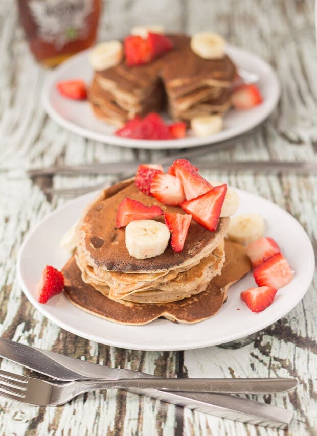 Two plates with a stack of strawberry banana pancakes on a plate topped with chopped strawberries. Knife and fork to the front. A bottle of maple syrup in the background.