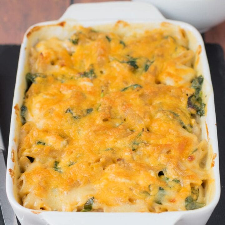 Easy chicken spinach pasta bake removed from oven. Sitting on a slate with a serving spoon to the side. Bowl of side salad in the background.