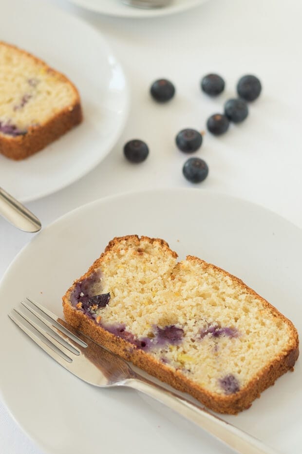 Close up of a slice of lemon blueberry yogurt cake on a plate with a fork. Another similar plate in the background with blueberries scattered around.