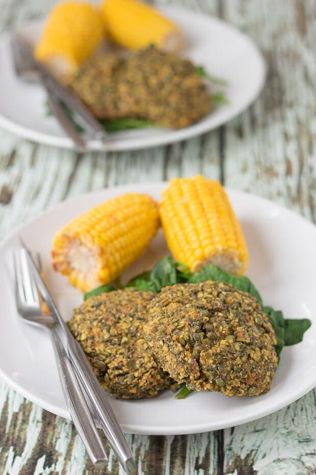 Two plates with two oven baked lentil and carrot patties on a plate with spinach and two corn on the cobs. Another similar plate in the background.