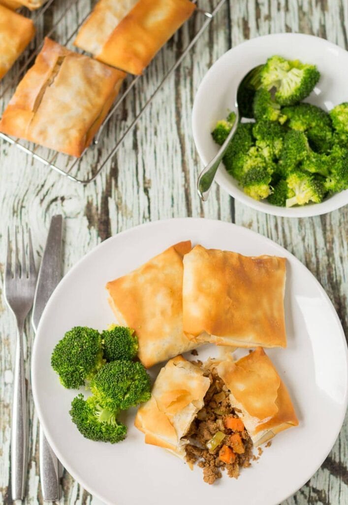 Birds eye view of a plate of three spiced quorn filo pastry parcels served on a plate with broccoli. One parcel cut in half to show filling. Fork to the left. Wire baking rack at top with more parcels on and a bowl of broccoli to the side.