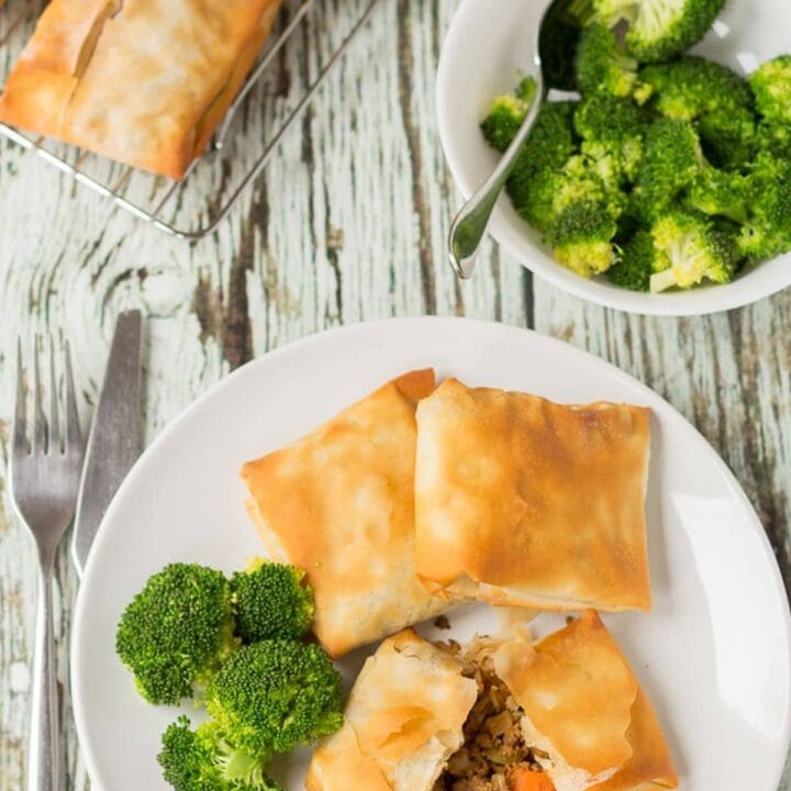 Birds eye view of a plate of three spiced quorn filo pastry parcels served on a plate with broccoli. One parcel cut in half to show filling. Fork to the left. Wire baking rack at top with more parcels on and a bowl of broccoli to the side.