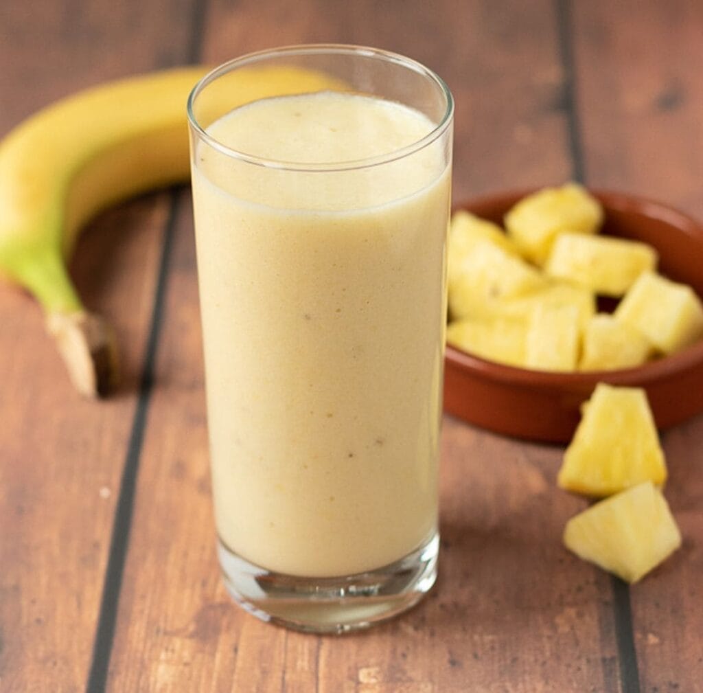 A glass of 4-ingredient tropical banana smoothie. A banana and a dish of pineapple cubes in the background.