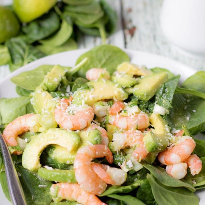 A plate of avocado prawn and coconut salad with a fork to the side. Some spinach leaves and a jug in the background as decoration.