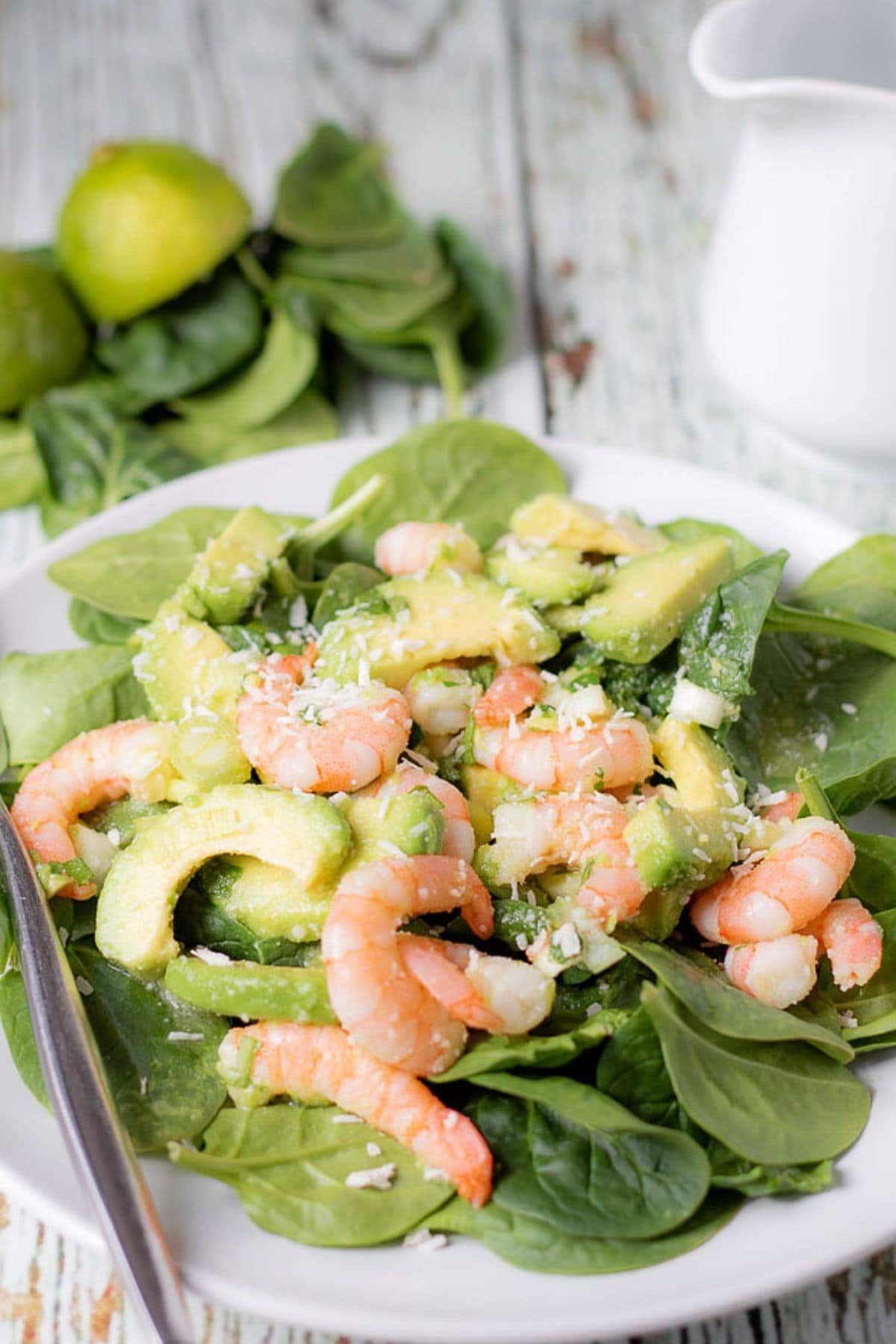 A plate of avocado prawn and coconut salad with a fork to the side. Some spinach leaves and a jug in the background as decoration.