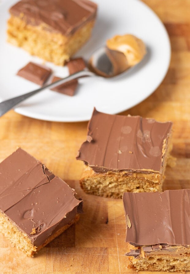 Three squares of chocolate peanut butter tray bake on a wooden chopping board with a square of chocolate peanut butter tray bake on a plate surrounded by squares of chocolate and a spoon with peanut butter on in the background.