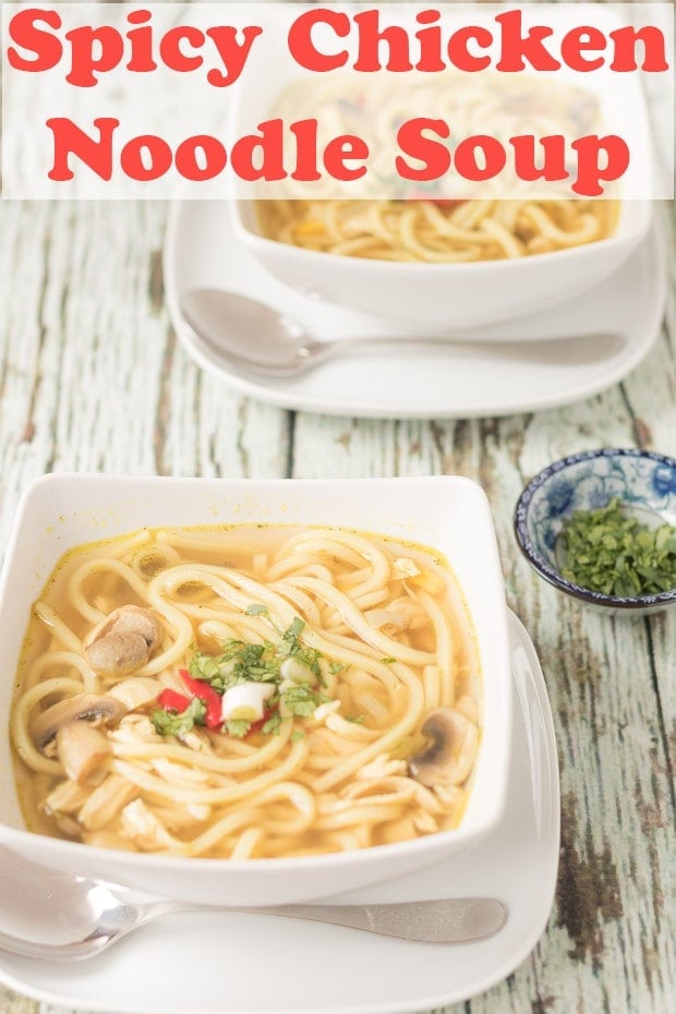 A square bowl of spicy chicken noodle soup sitting on a square plate with a soup spoon in front. A dish of chopped coriander as garnish to the right side. Pin title text overlay at top.
