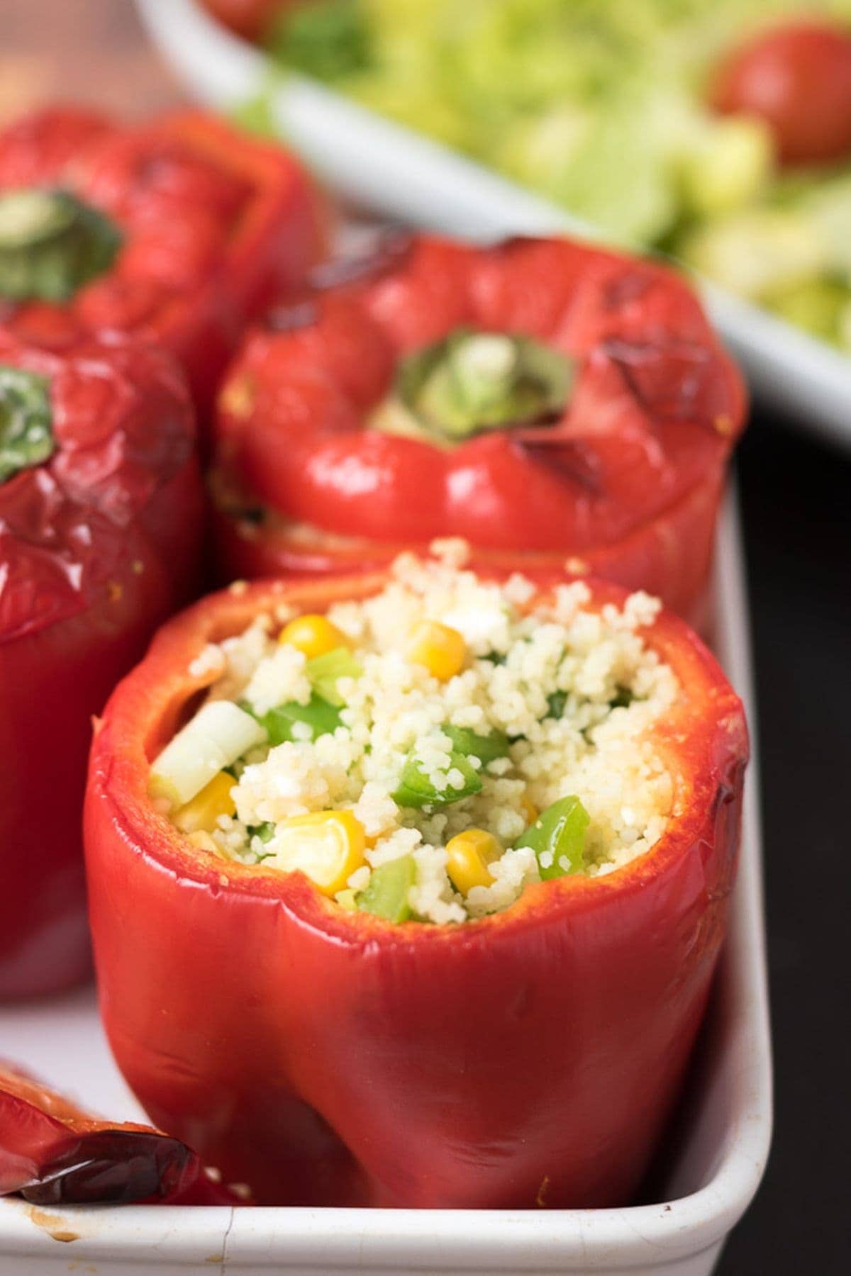 Close up of vegetarian couscous stuffed peppers sitting upright in a casserole dish showing the filling of the front pepper. Salad dish in the background.