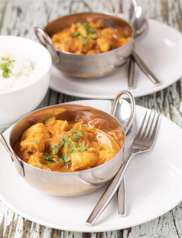Close up of two balti dishes sitting on plates served with chicken sharabi curry in. Knife and fork to the side and bowl of rice in between.