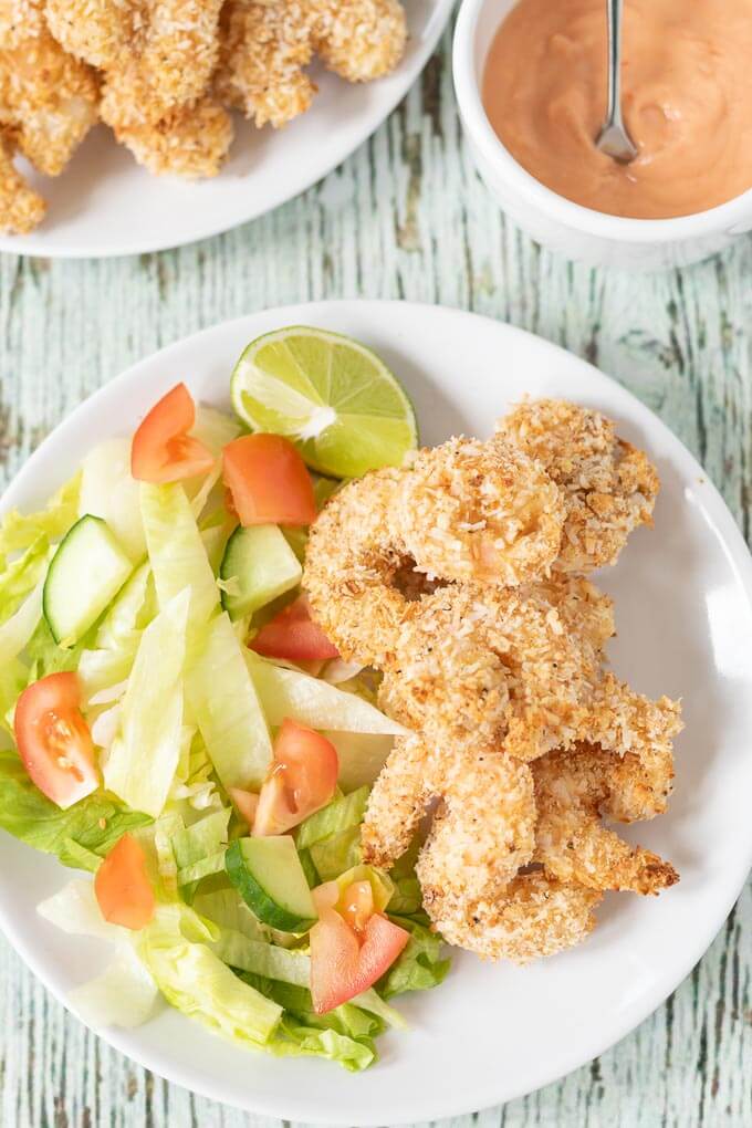 Birds eye view of a plate of healthy baked coconut prawns served with salad and a lime wedge. Dipping sauce and another plate of baked prawns above.