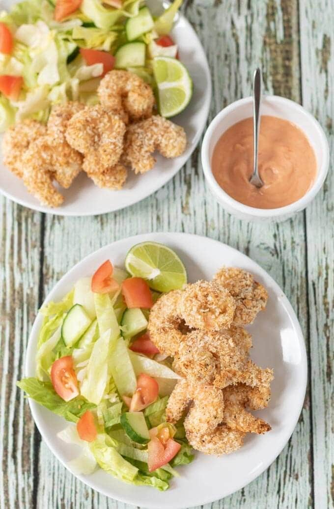 Birds eye view of two plates of healthy baked coconut prawns served with salad and a lime wedge. Dipping sauce in between.