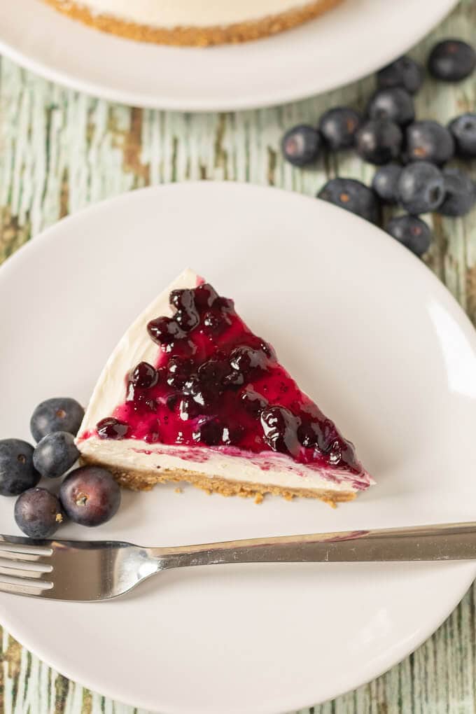 A slice of no bake blueberry cheesecake on a plate with a fork and some blueberries beside.