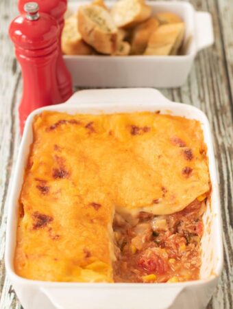 A casserole dish of tuna sweetcorn lasagne with a portion removed. Salt and pepper cellars and a bowl of garlic bread in the background.