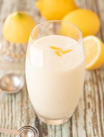 A glass of zingy lemon smoothie with measuring spoons at the base and three lemons in the background as decoration.