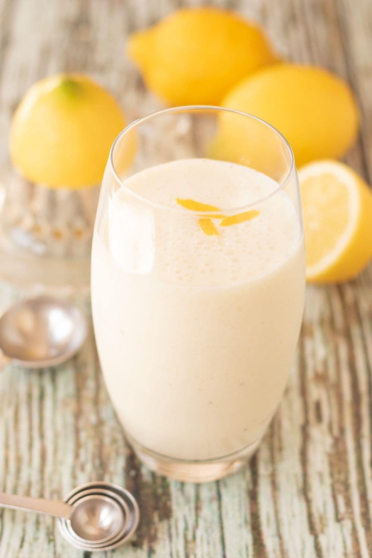 A glass of zingy lemon smoothie with measuring spoons at the base and three lemons in the background as decoration.