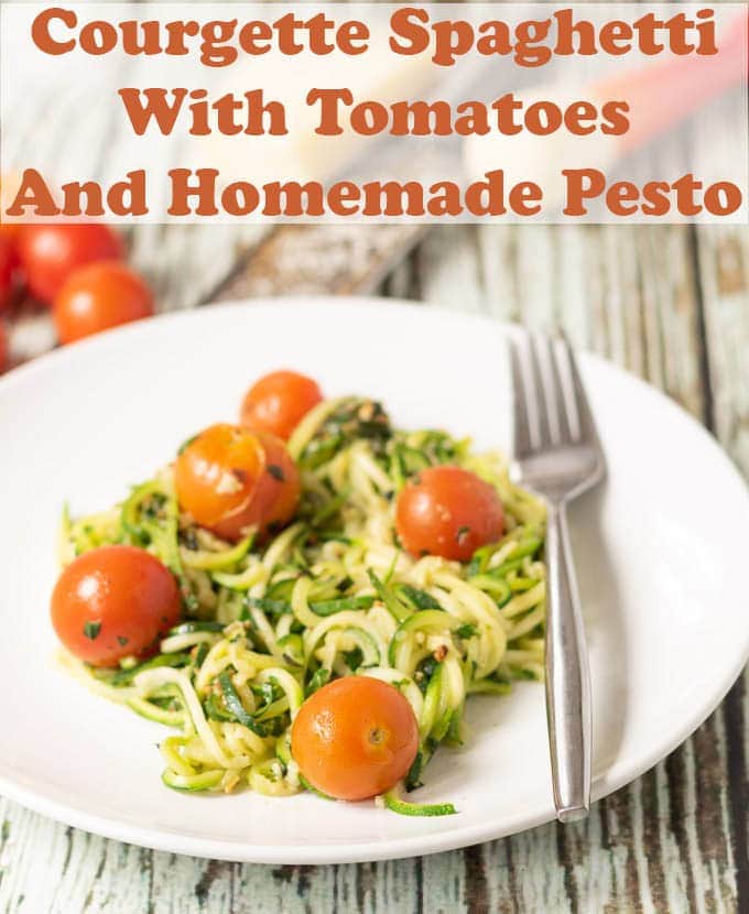 Courgette spaghetti with tomatoes and homemade pesto served on a white plate with a fork ready to eat. Cherry tomatoes in the background as decoration.