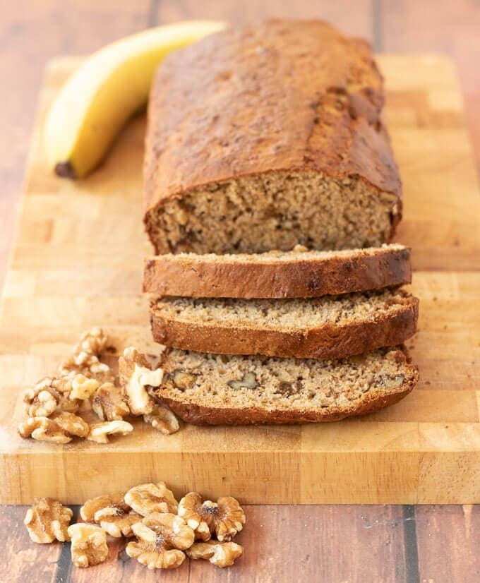 Healthy banana and walnut loaf on a chopping board. Three slices cut off at the front. A banana in the background and walnuts in the foreground.