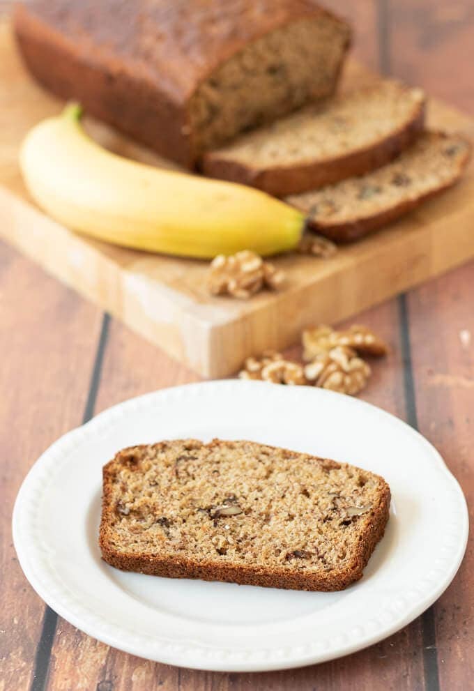 A slice of healthy banana and walnut loaf on a plate. Rest of the loaf on a chopping board. Three slices cut off and a banana and walnuts surrounding as decoration in the background.