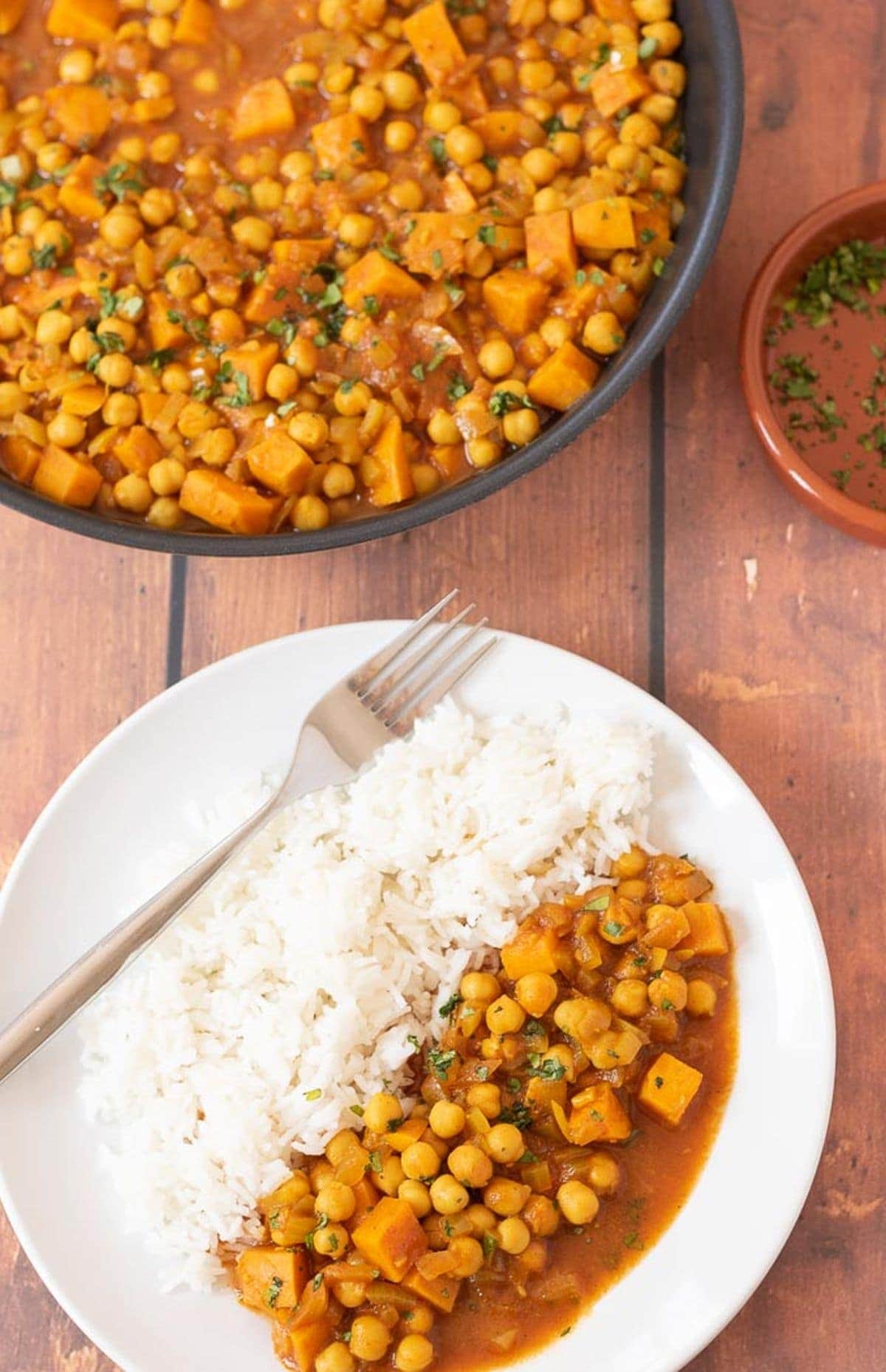 Birds eye view of a plate of sweet potato and chickpea curry served with rice and a fork to the side. Rest of the curry in a pan at the top with a dish of garnish to the side.