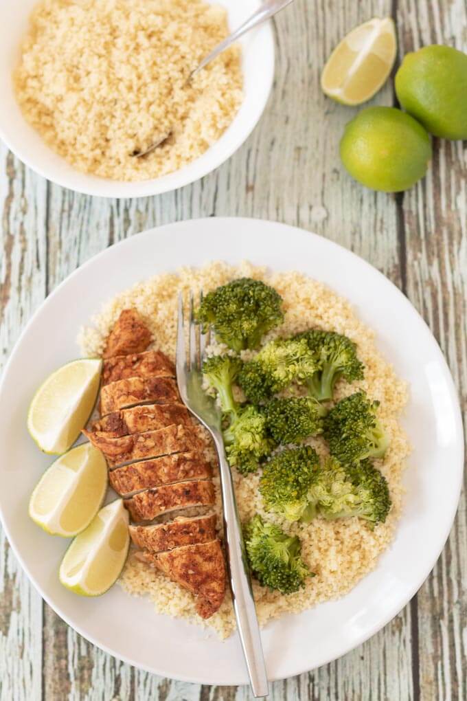 Birds eye view of a plate with oven baked lime chicken served on a bed of rice and broccoli with three lime wedges beside. A bowl of rice and two and a half limes in the background.