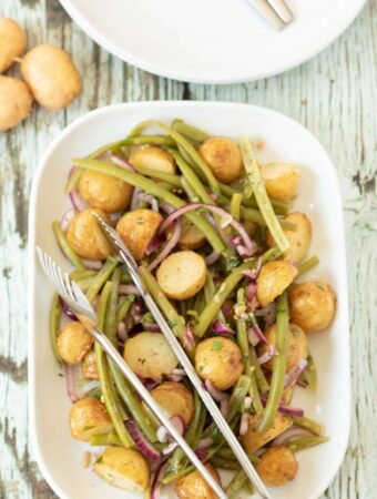 Birds eye view of roast potato and green bean salad served on a platter with serving tongs on top. Part of a plate and three potatoes at the top.