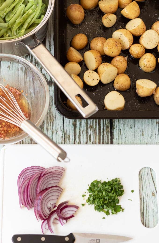 Birds eye view of a chopping board with a knife and chopped red onion and parsley on. The vinaigrette in a bowl with a whisk, part of a saucepan with green beans in and the baking tray with potatoes in.