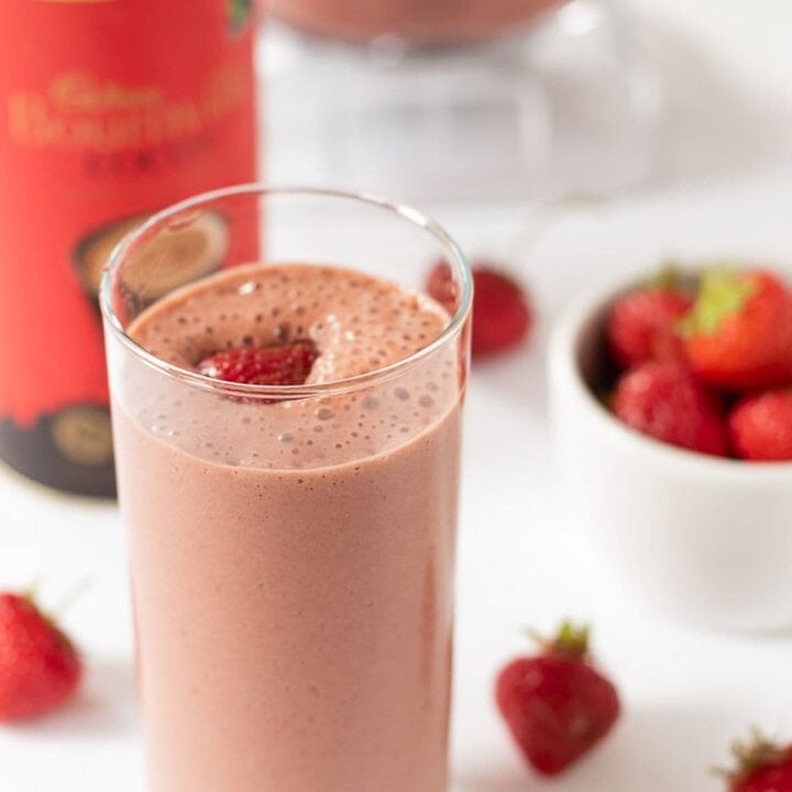 A tall glass of easy chocolate strawberry smoothie. Container of drinking chocolate, a dish of strawberries to the side and the rest of the smoothie in a blender jug in the background.