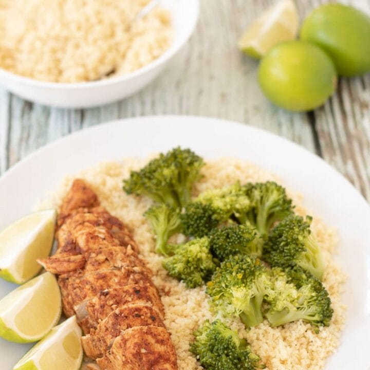 A plate with oven baked lime chicken served on a bed of rice and broccoli with lime wedges beside. A bowl of rice and two and a half limes in the background.