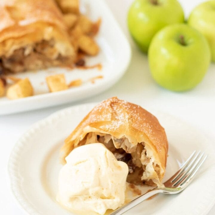 Sugar free easy apple strudel served on a plate with a blob of ice cream and a fork beside. Rest of the strudel and three apples in the background.